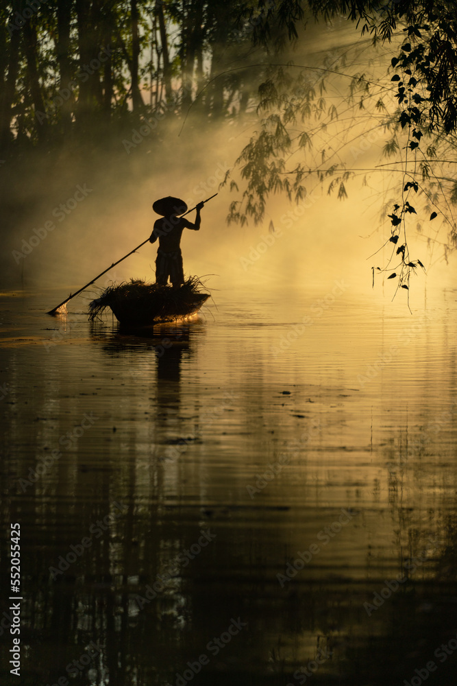 Photo of a local asian old man male boatman wearing conical hat rowing a small wooden boat across a small river during sunset time in a bamboo forest to deliver some dry grasses as animal feeds. 