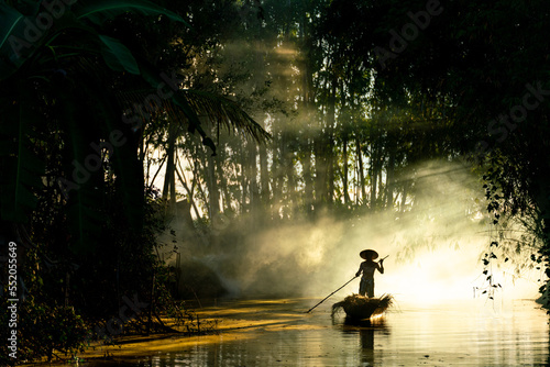 Photo of a local asian old man male boatman wearing conical hat rowing a small wooden boat across a small river during sunset time in a bamboo forest to deliver some dry grasses as animal feeds. 