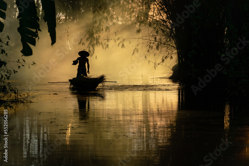 Photo of a local asian old man male boatman wearing conical hat rowing a small wooden boat across a small river during sunset time in a bamboo forest to deliver some dry grasses as animal feeds. 