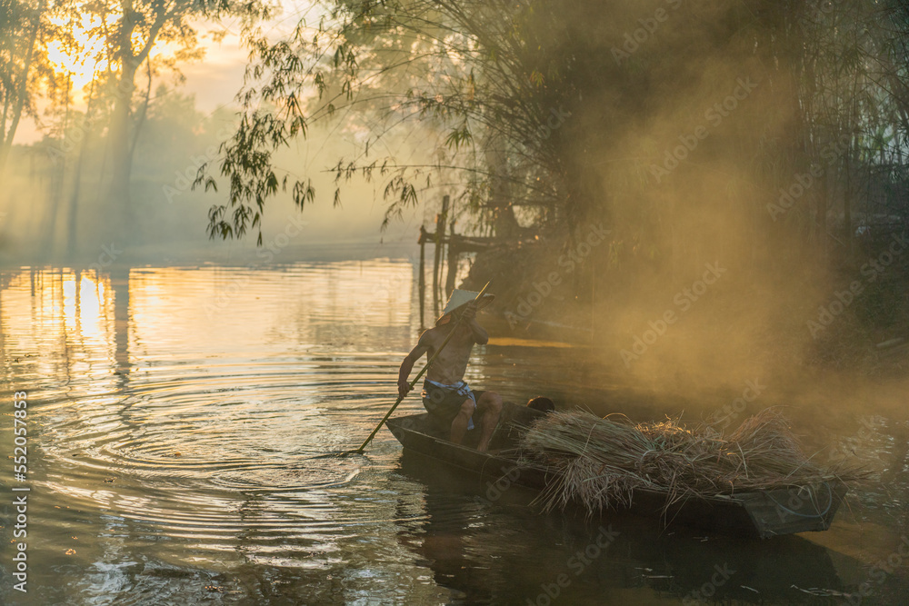 Φωτογραφία Photo of a local asian old man male boatman wearing conical hat rowing a small wooden boat across a small river during sunset time in a bamboo forest to deliver some dry grasses as animal feeds