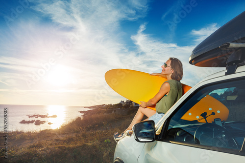 Woman with surfboard sit on a car hood look over sea and sunset