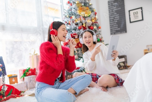 Two young asian female lady friends in a room listening to new songs on their headphone via their digital tablet celebrate Christmas together