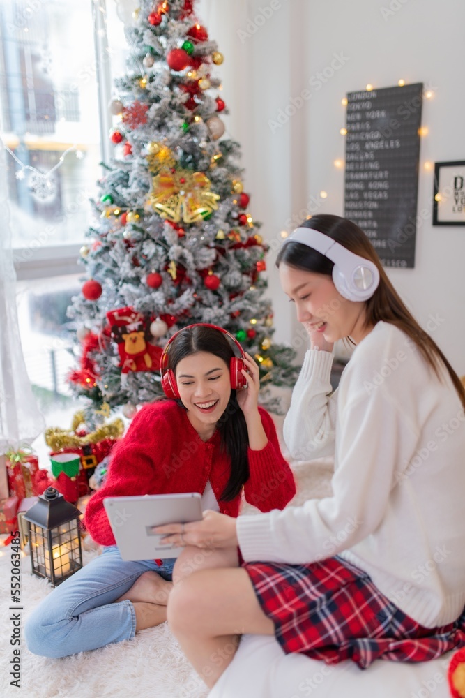 Two young asian female lady friends in a room listening to new songs on their headphone via their digital tablet celebrate Christmas together Two young asian female lady friends in a room listening to new songs on their headphone via their digital tablet celebrate Christmas together