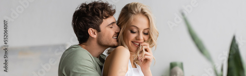 Positive man in pajama kissing young girlfriend at home in morning, banner.