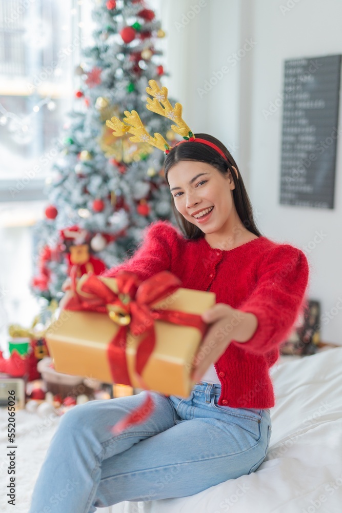 Cute beautiful young asian lady woman wearing reindeer headband holding a gold color gift box with red ribbon posing in front a big Christmas tree with lots of decoration lights gift box and ornaments Cute beautiful young asian lady woman wearing reindeer headband holding a gold color gift box with red ribbon posing in front a big Christmas tree with lots of decoration lights gift box and ornaments