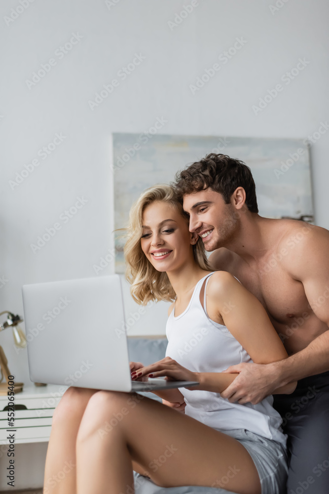 Shirtless man hugging blonde girlfriend with laptop in bedroom.