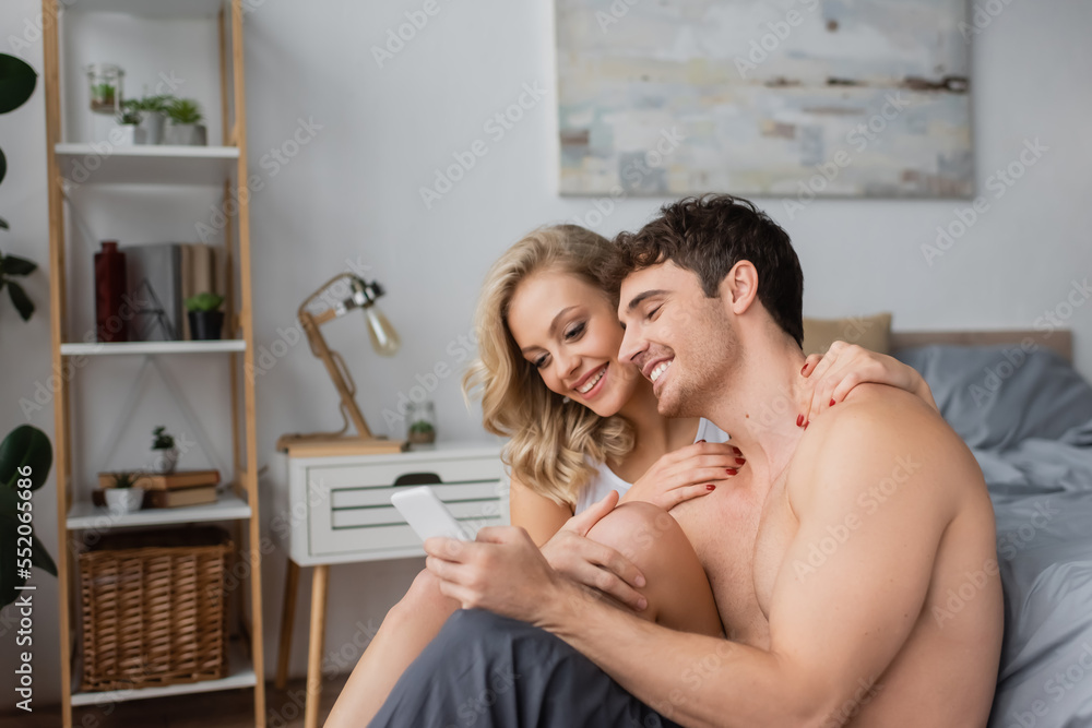 Smiling woman hugging sexy boyfriend with cellphone in bedroom. Smiling woman hugging sexy boyfriend with cellphone in bedroom.