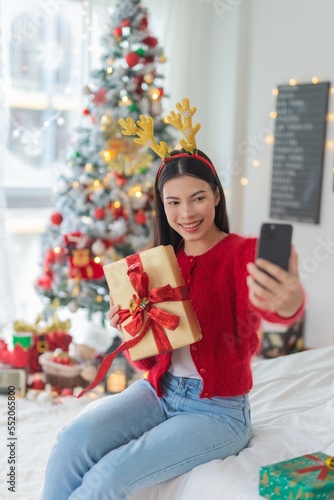 Cute beautiful young asian lady woman wearing reindeer headband selfie with her mobile phone holding gift box posing in front a big Christmas tree with lots of decoration lights gift box and ornaments © asean studio