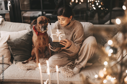 Happy woman with dog cuddling and unpacking gifts, looking excitedly into the box, in a cozy environment at home on the couch, mysterious, warm colors