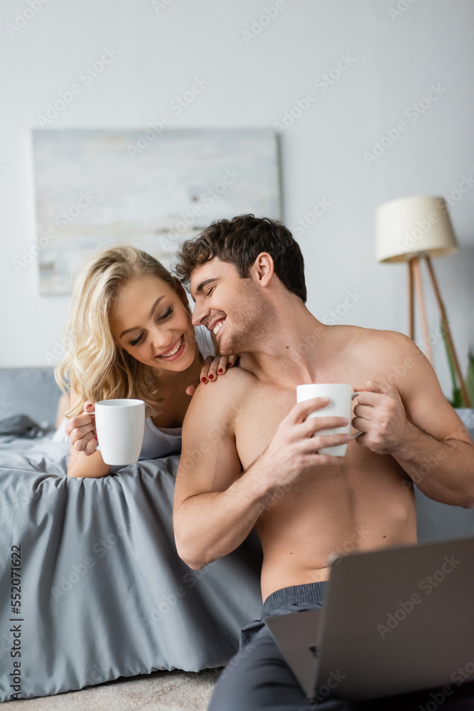 Smiling shirtless man holding cup of coffee near blonde girlfriend and laptop in bedroom. Smiling shirtless man holding cup of coffee near blonde girlfriend and laptop in bedroom.