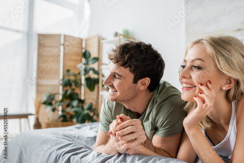 Happy man in pajama holding hand of blonde girlfriend on bed in morning.