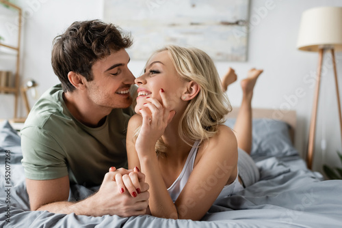 Smiling man in pajama kissing and holding hand of cheerful girlfriend on bed.