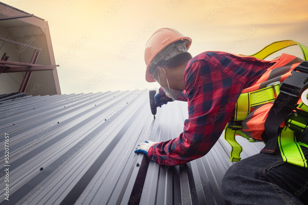 Roofer worker in protective uniform wear and gloves,Roofing tools,installing new roofs under construction,Electric drill used on new roofs with metal sheet. Roofer worker in protective uniform wear and gloves,Roofing tools,installing new roofs under construction,Electric drill used on new roofs with metal sheet.
