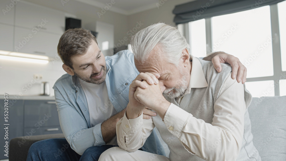 Fototapeta premium Son supporting upset senior father, family sitting on sofa at home, care