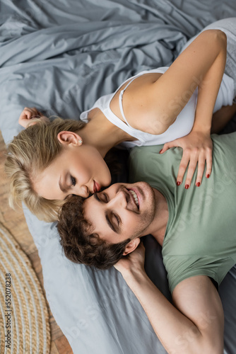 Top view of young woman kissing cheerful boyfriend in pajama on bed at home.