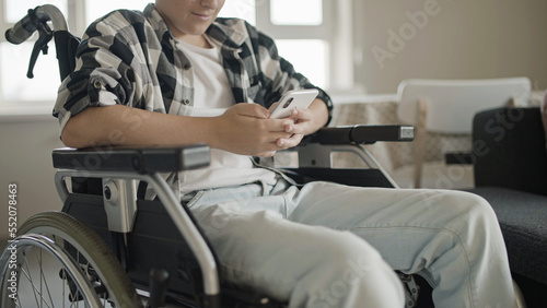 Teenage boy in wheelchair sending message on smartphone, texting with friends, close-up