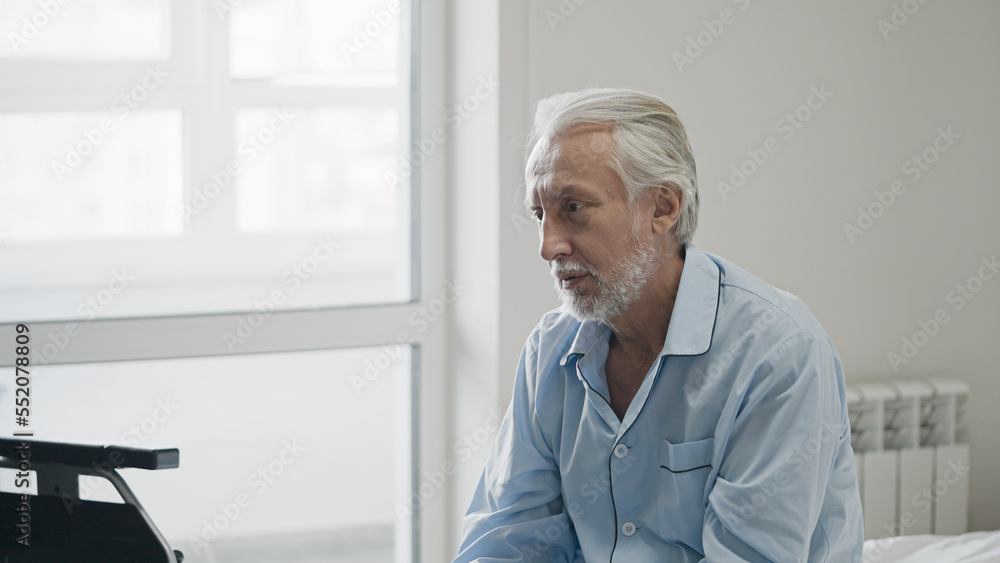 Sad old man with disability sitting on bed and looking at his wheelchair