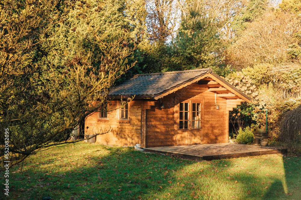 Fototapeta premium Small wooden house in a beautiful garden with lots of sun. Garden in summer or spring. Wooden garden shed. Secluded little cottage in the woods