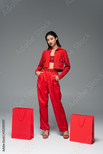 young asian model in red jacket and trousers posing with hands in pockets near shopping bags on grey background.