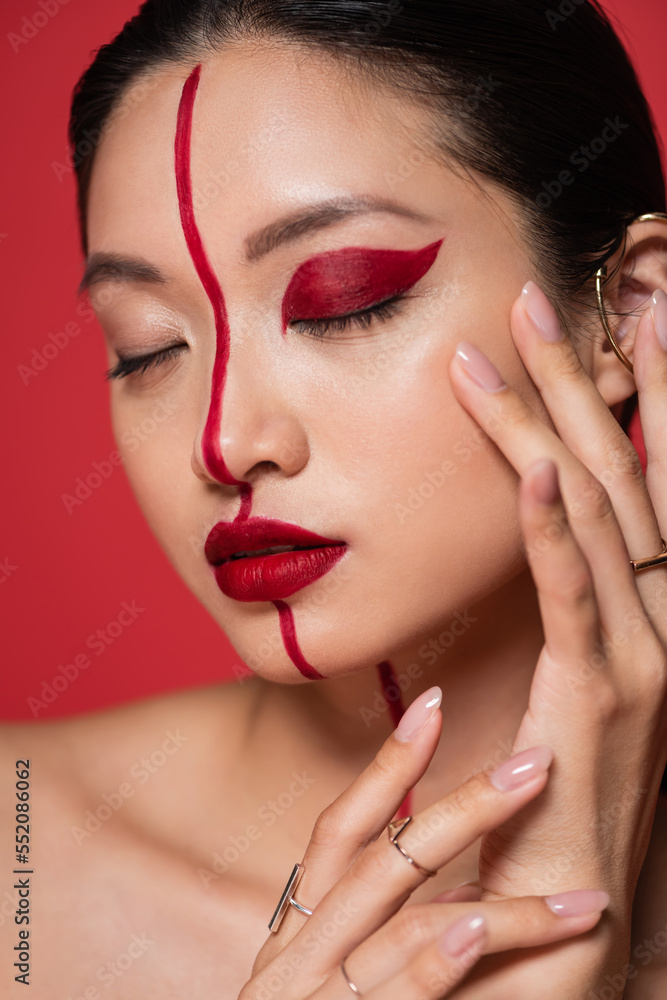 Fototapeta premium close up portrait of asian woman with closed eyes and creative makeup touching face isolated on red.