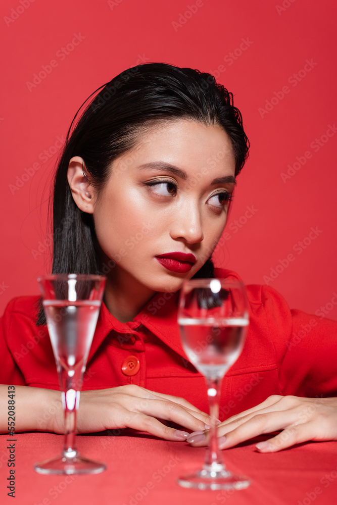 portrait of brunette asian woman with makeup looking away near blurred glasses of water isolated on red. portrait of brunette asian woman with makeup looking away near blurred glasses of water isolated on red.