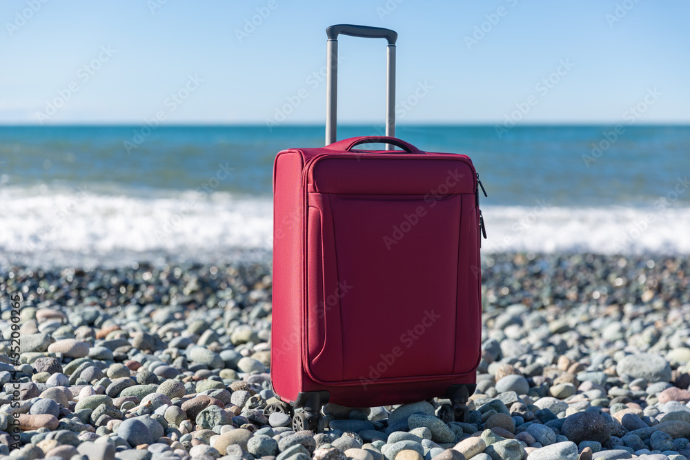 Color travel suitcase on pebble beach with turquoise sea background, summer holidays concept