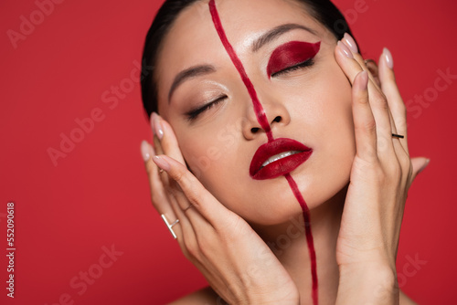 portrait of sensual asian woman touching perfect face with bright artistic makeup while posing with closed eyes isolated on red.