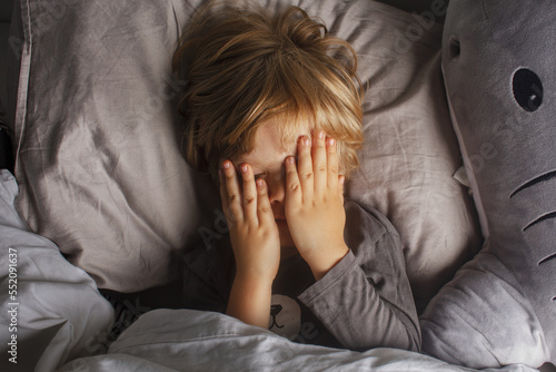 The child was scared before going to bed. Night terrors in a child. The kid covers his face with his hands in a fear. Children's experiences. Boy in the bed. View from above. Sad psychological state. 
