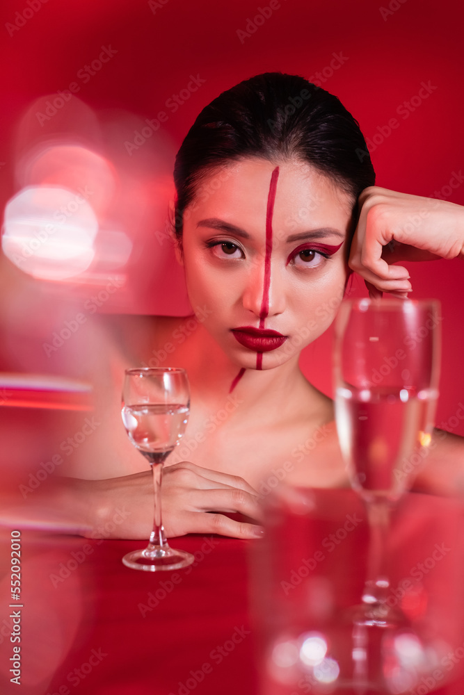 portrait of asian woman with makeup on face divided with line looking at camera near blurred glasses on red background. portrait of asian woman with makeup on face divided with line looking at camera near blurred glasses on red background.