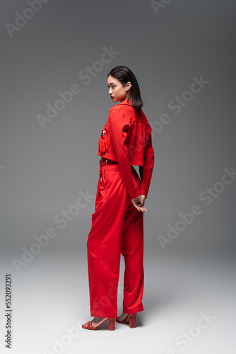 full length of asian woman in red blazer and pants standing on grey background.