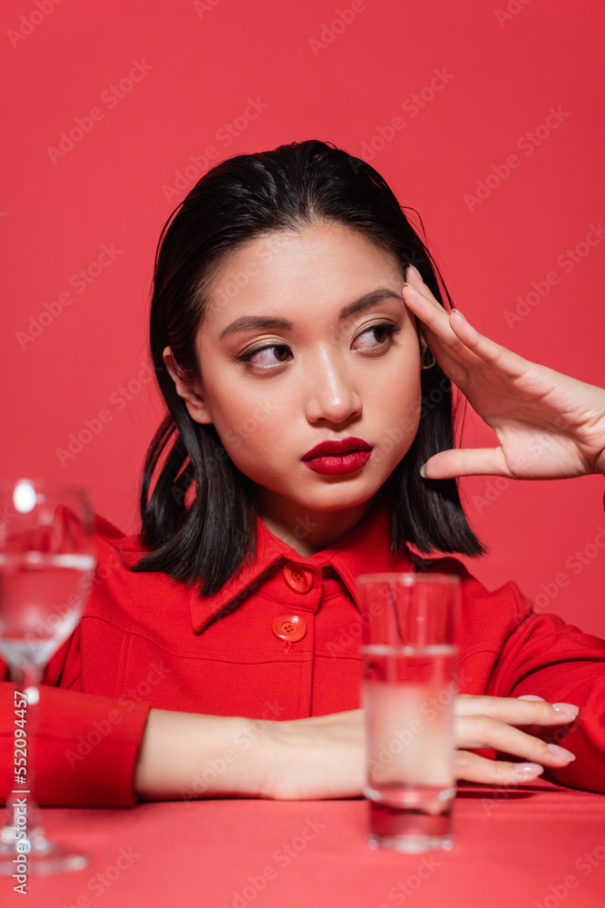 stylish asian woman with makeup holding hand near face and looking away near blurred glasses with water isolated on red. stylish asian woman with makeup holding hand near face and looking away near blurred glasses with water isolated on red.