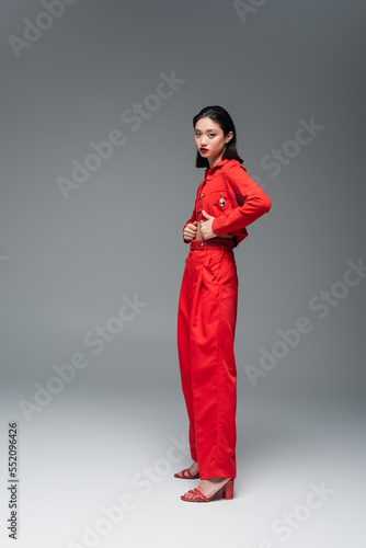 full length of young asian woman in red jacket and trousers looking at camera on grey background.