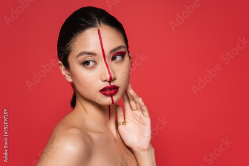 pretty asian woman with naked shoulder and artistic visage touching face while looking away isolated on red.