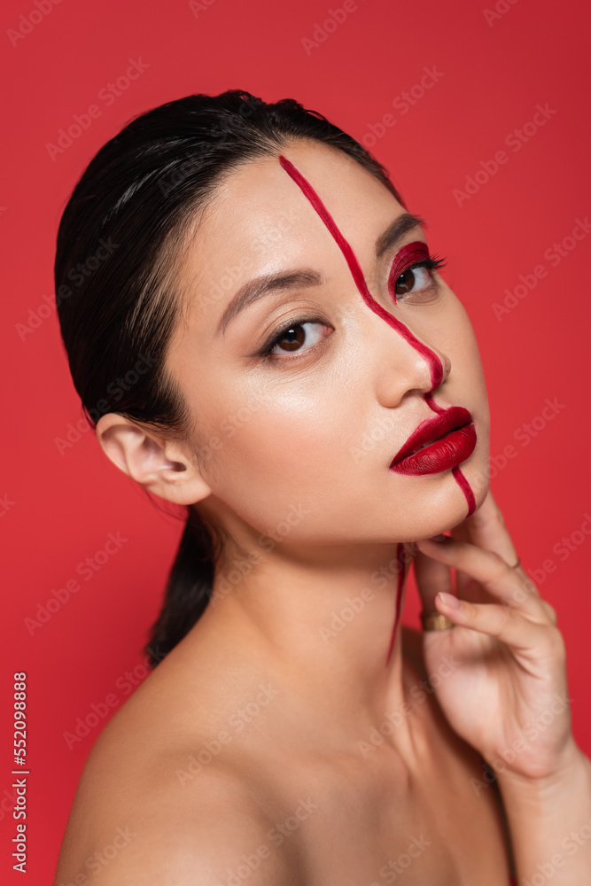 Fototapeta premium portrait of asian model with creative visage and perfect skin holding hand near face and looking at camera isolated on red.