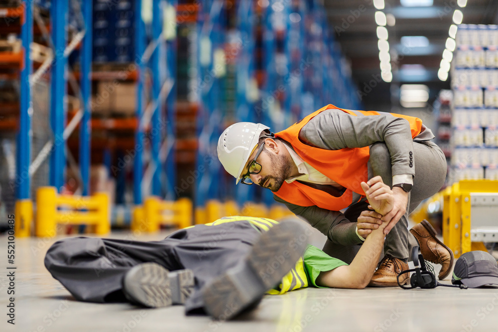 A warehouse worker is lying on the ground unconscious and his supervisor is giving him first aid. A warehouse worker is lying on the ground unconscious and his supervisor is giving him first aid.