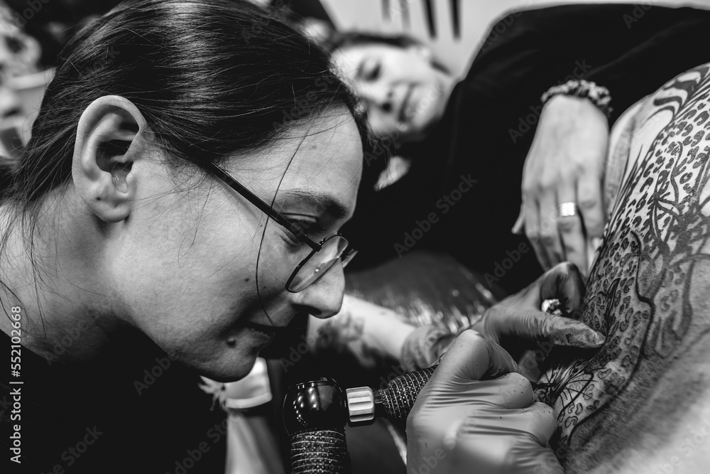 Smiling young tattoo artist girl with glasses making a tattoo, with tattoo machine, of a big tiger, in the thigh and hips of a happy and beautiful woman (in black and white)