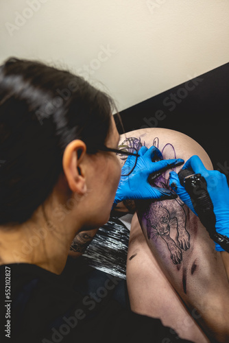 Young tattoo artist girl with glasses making a tattoo, with tattoo machine, of a big tiger, in the thigh and hips of a beautiful woman photo