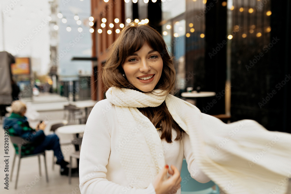 Excited lovely pretty girl with wonderful smile having outdoor on background of city cafe with lights. She is playing with her scarf and dancing
