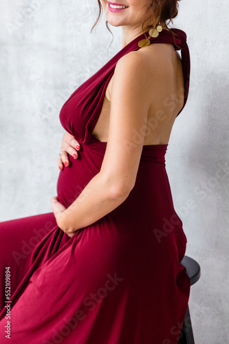 A pregnant woman in a long burgundy silk dress hugs her belly and poses on a gray background, sitting on a chair