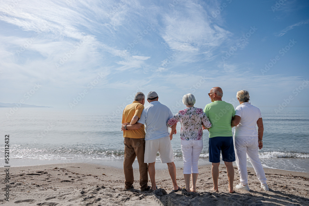 gruppo di 5 anziani in spiaggia in piedi guardano abbracciati l'orizzonte in lontananza gruppo di 5 anziani in spiaggia in piedi guardano abbracciati l'orizzonte in lontananza