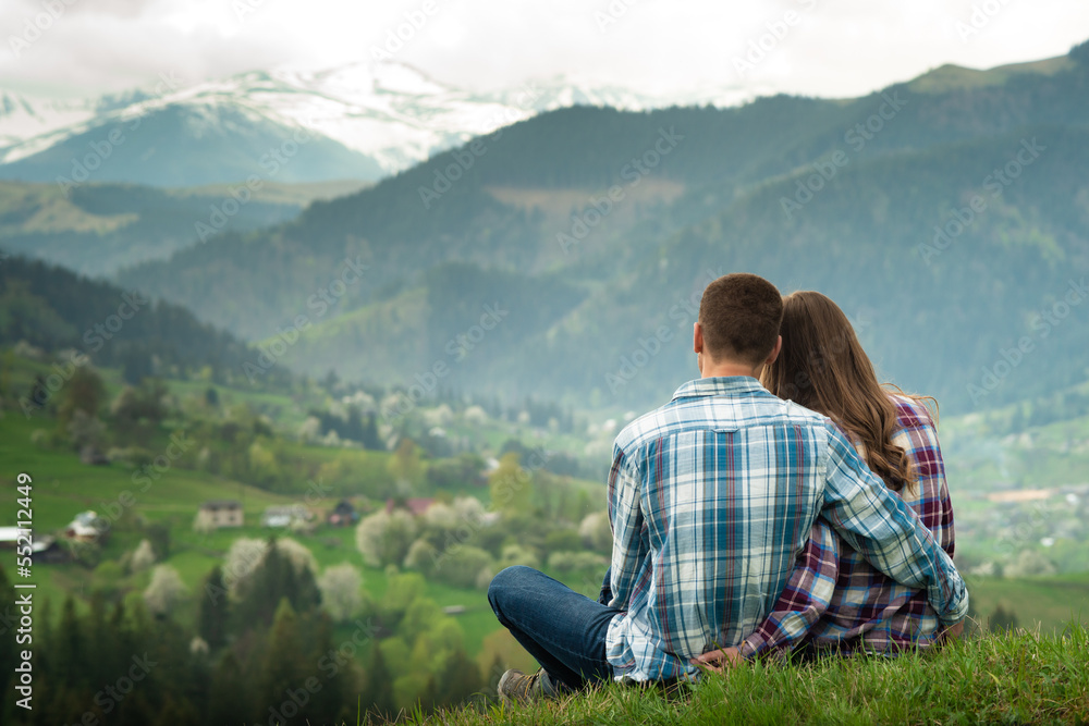 A couple in love sit in embraces on the top of mountain with a scenic view on the background. Side view