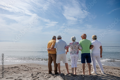 gruppo di 5 anziani in spiaggia in piedi guardano abbracciati l'orizzonte in lontananza 