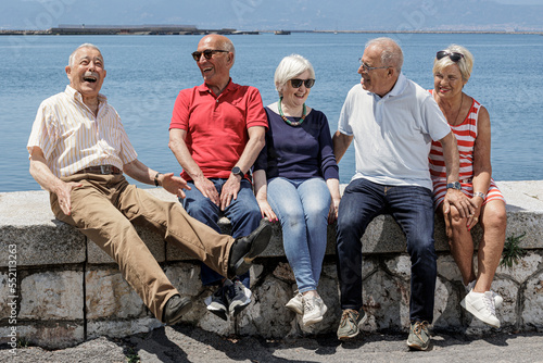gruppo di anziani amici seduti nel muretto di un porto di mare, si rilassano chiacchierando felici.