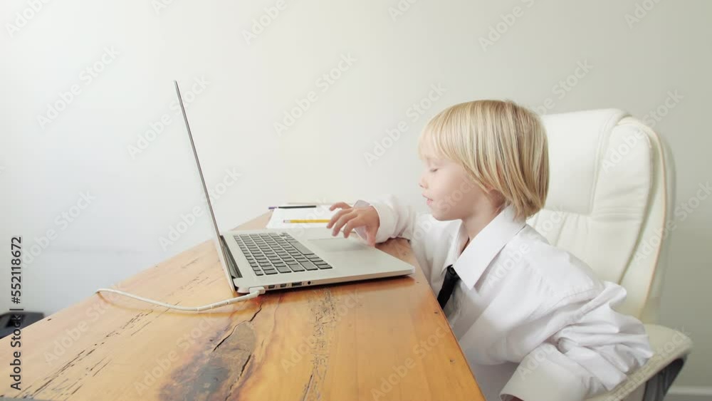 Cute caucasian 5 years old boy in his fathers's shirt and tie is looking at the laptop screen and pressing buttons pretending to be an adult. Home office. Funny moment Cute caucasian 5 years old boy in his fathers's shirt and tie is looking at the laptop screen and pressing buttons pretending to be an adult. Home office. Funny moment