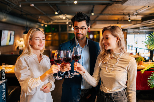 Portrait of elegant man in suit and eyeglasses and two pretty blonde women holding glasses of red wine, standing posing in restaurant. Happy young male and female enjoying nice dinner.