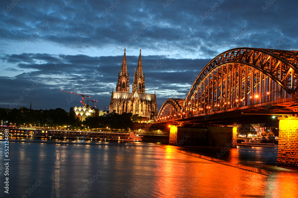 Fototapeta premium Cologne, Germany. Famous Hohenzollern Bridge over Rhine river. Buildings in historic city centre. Towers of Cathedral and city hall by night. 