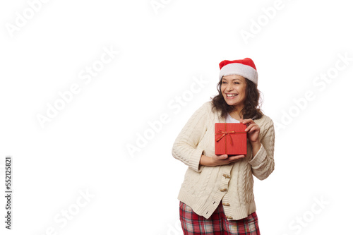 Waist-up portrait on white background of a multi-ethnic positive pretty woman wearing Santa hat, cheerfully smiling looking aside at copy ad space, posing with gift box with Christmas happy present