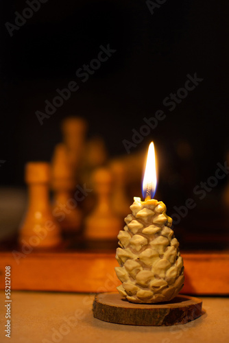 Burning candle in pine cone shape. Chess in the background. Handmade. Holiday advent