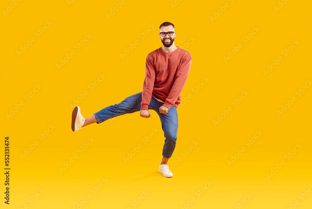 Smiling funky cheerful young bearded man with short hair in glasses wearing red sweatshirt and denim pants dancing on one leg isolated on yellow background. Good mood happy lifestyle nice day concept.