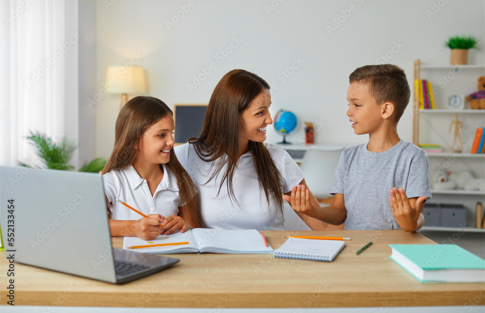 Caring and friendly young mother helps her children to complete their school homework. Woman with her daughter and son are talking and writing while sitting at table in front of laptop. Caring and friendly young mother helps her children to complete their school homework. Woman with her daughter and son are talking and writing while sitting at table in front of laptop.
